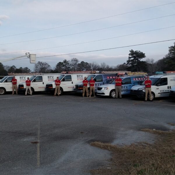 A group of men in red shirts stand in front of a line of branded service vehicles in an open parking lot. The vehicles include vans, trucks, and a bucket truck, all with the company logo 