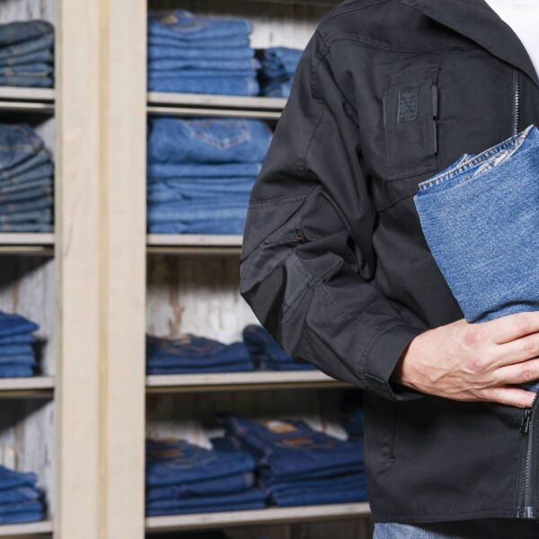 A person in a black jacket concealing a pair of folded blue jeans inside their coat. Behind them, shelves are filled with neatly folded and stacked jeans. They appear to be in a clothing store.