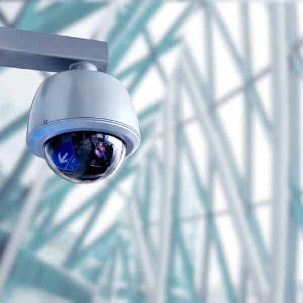 A close-up of a security camera mounted on a wall inside a modern building. The camera is housed in a dome enclosure, and the background features large windows with a sleek, futuristic architectural design, characterized by steel beams and glass panels.