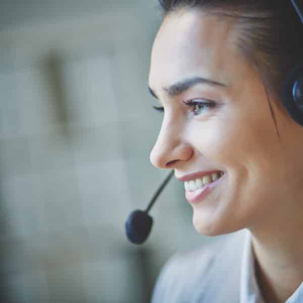 A person with dark hair is smiling and wearing a headset with a microphone. The individual appears to be engaging in a conversation, possibly providing customer service or technical support. The background is blurred, focusing attention on the person.