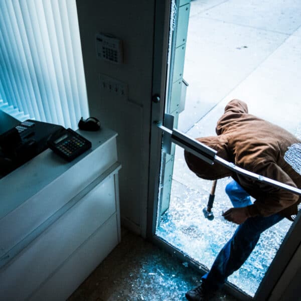A person wearing a brown hooded jacket and jeans is breaking into a storefront with a hammer, causing shattered glass around the door. The interior shows a cash register on the counter and some shelves with colorful bowls. The outside sidewalk is visible.