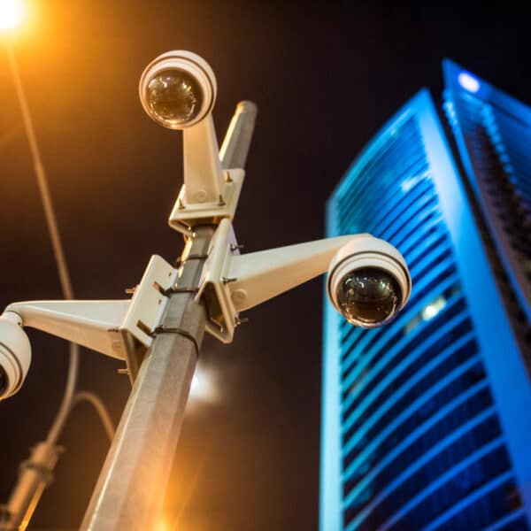 A view of several security cameras mounted on a pole, with a high-rise building illuminated in blue lights in the background. The scene is captured at night, with a street lamp casting an orange glow nearby.