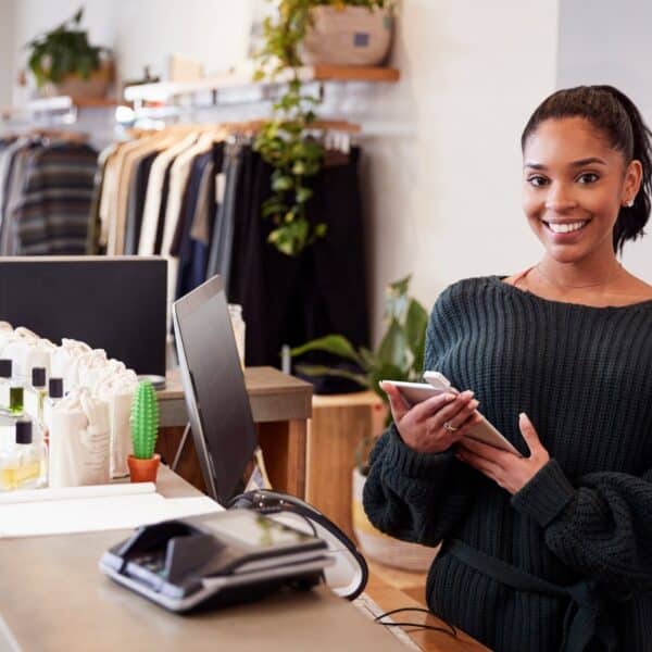 A retail worker at a clothing store stands behind the counter holding a tablet and smiling. The counter has a payment terminal, a potted cactus, and some bottles of products. In the background, clothing racks and green plants are visible.