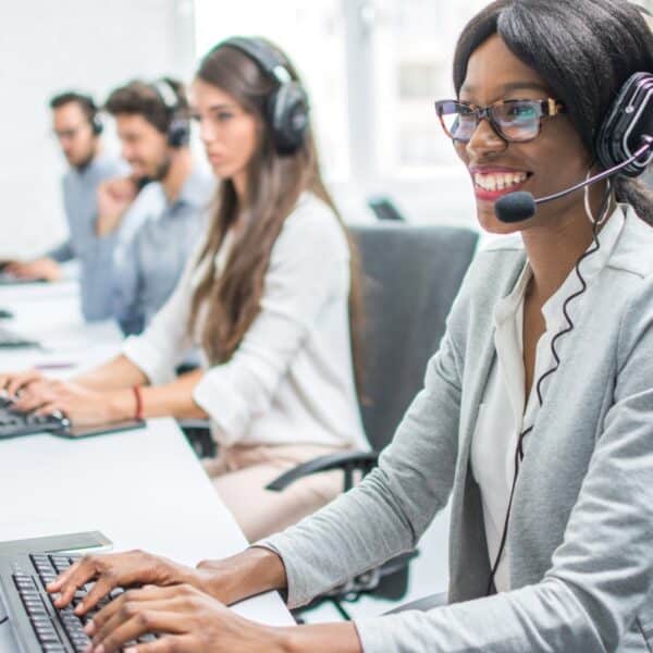 A group of customer service representatives working in an open-plan office. The focus is on a smiling woman wearing glasses and a headset, typing on a keyboard. Three colleagues, also wearing headsets, are working at computers in the background.