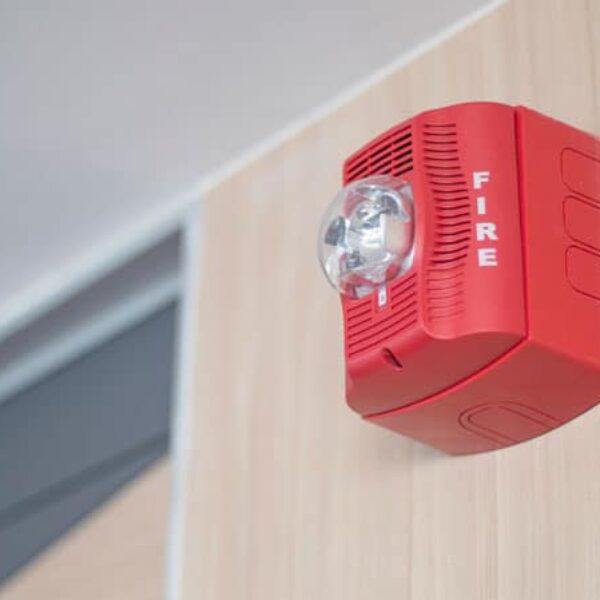 A red fire alarm device is mounted on a light wooden wall. The alarm features a clear strobe light and has the word 