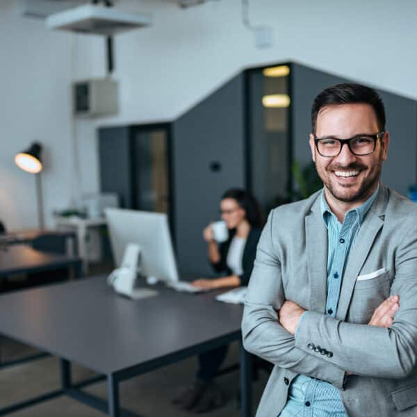 A man in a light gray suit and glasses stands smiling with his arms crossed in a modern office. Behind him, a woman works at a computer and drinks from a mug. The office features minimalist decor with large desks and a lamp.