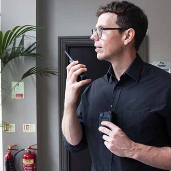 A man in a black shirt uses a walkie-talkie while standing indoors. He is near a large green plant and a pair of red fire extinguishers. He holds a small device in his other hand and looks to the right. The background shows a gray wall and a door.