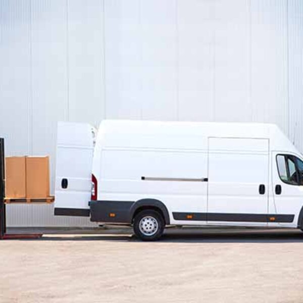 A worker operates an electric pallet jack to load large cardboard boxes into a white delivery van parked against a building with a corrugated metal wall. The van’s side door is open, and the worker is guiding the pallet towards the vehicle.