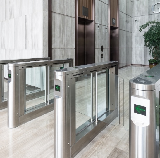 An indoor security checkpoint with three metallic turnstiles with glass barriers. Card readers with green lights are on top of each turnstile. An elevator lobby with two elevators is visible in the background, and a potted plant is on the right side.