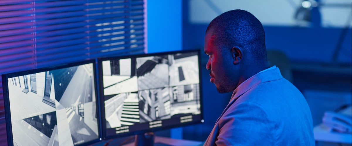 A person wearing a blazer is monitoring multiple security camera feeds on two computer screens in a dimly lit room with blue lighting. The screens display various indoor and outdoor scenes from different cameras. Blinds cover a window to the left.