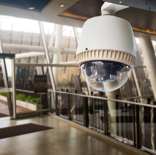 A white security camera dome is mounted on the ceiling of a modern, glass-walled building with plants and railings in the background. The camera lens is visible inside the transparent dome, and the setting appears to be a public or commercial space.