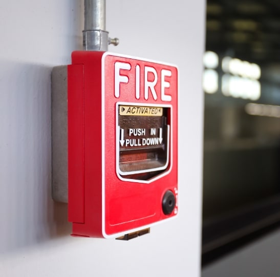 A red fire alarm is mounted on a white wall with a metallic pipe connected at the top. The alarm reads "FIRE" in large white letters, with instructions to "PUSH IN PULL DOWN" below. A small black circular element is located at the bottom right.