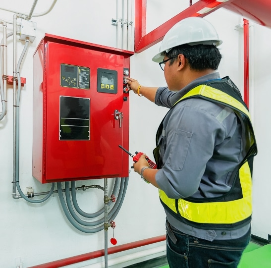 A worker wearing a white hard hat, safety vest, and glasses is adjusting a control panel on a red, wall-mounted electrical box. He is holding a walkie-talkie and working in an industrial environment with visible cables and pipes.