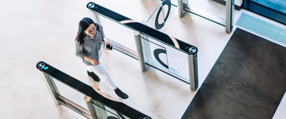 A woman walking through a security gate in a bright building. She is holding a phone and wearing a striped shirt and white pants. The gate has a clear barrier and green lights indicating access. The floor is tiled, and a dark mat is placed near the entrance.