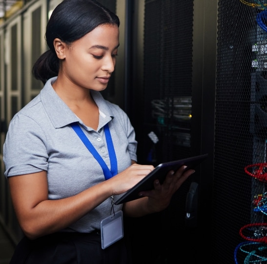 A person in a gray polo shirt and blue lanyard works on a tablet in a server room. They are standing next to a server rack with various cables visible. The environment appears to be professional and focused on technology.