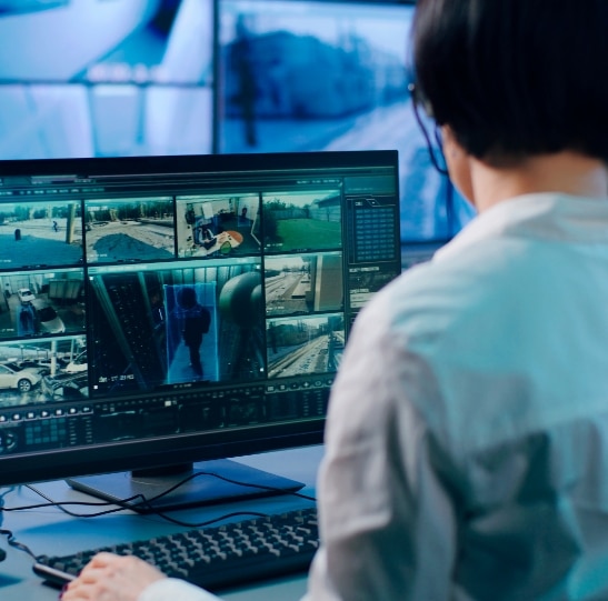A person is sitting at a desk, monitoring multiple security camera feeds displayed on a large computer screen. The screen shows various surveillance footage, including outdoor and indoor scenes. The setting appears to be a surveillance control room.